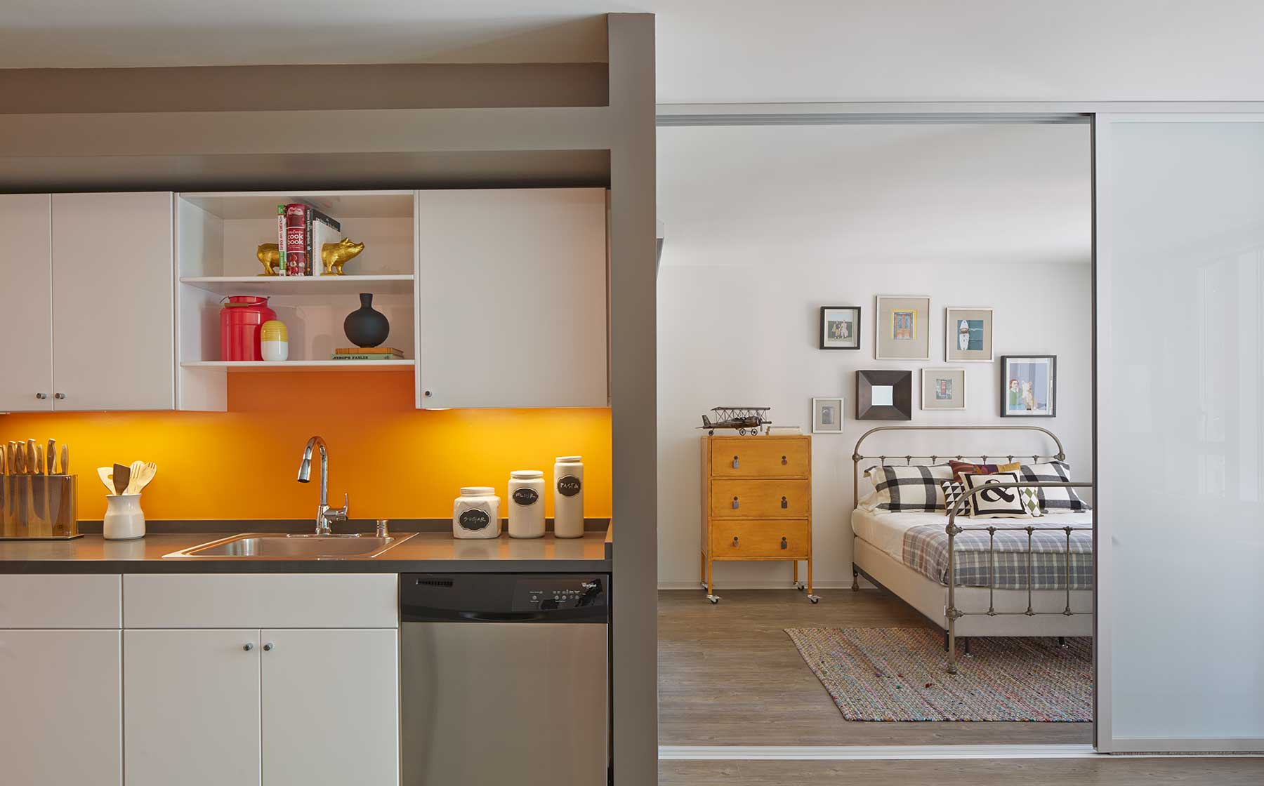 Open concept kitchen with white cabinetry, dark grey countertops with color backsplash stainless steel appliances and bedrrom with sliding barn door and hard surface flooring